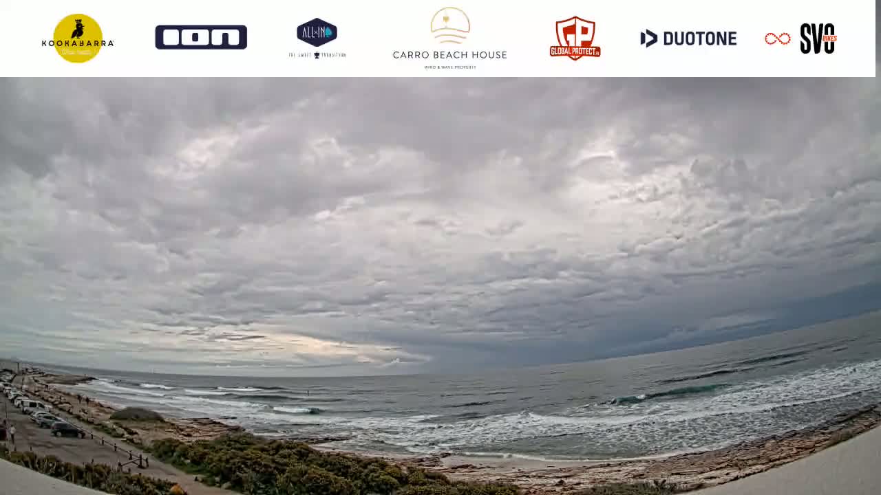 An overcast sky hangs over a beach with gently rolling waves and a rocky shoreline, with vegetation visible along the coast.