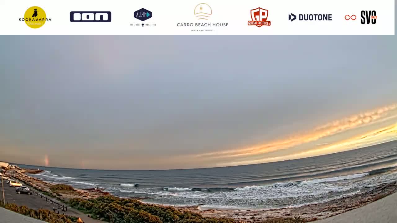 A wide shot of an ocean beach at sunset, with small waves and a mostly cloudy sky showing hints of orange and yellow.