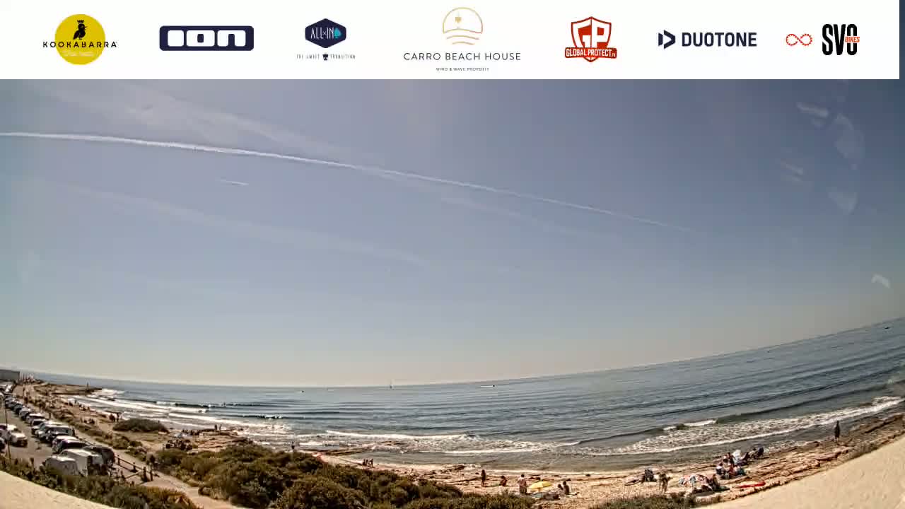 A wide shot shows a sandy beach with people relaxing and a calm ocean under a clear, sunny sky.