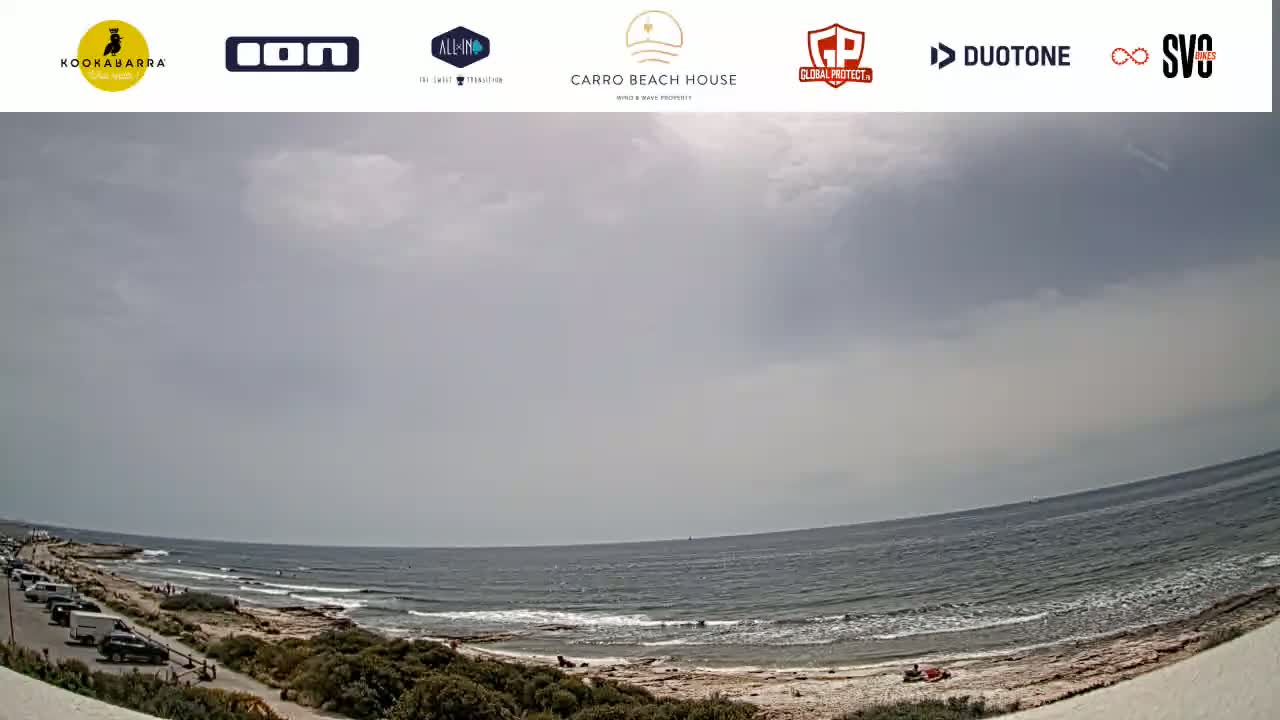 A mostly cloudy day at a beach with a rocky shoreline and a few people visible, some cars parked in a lot nearby.