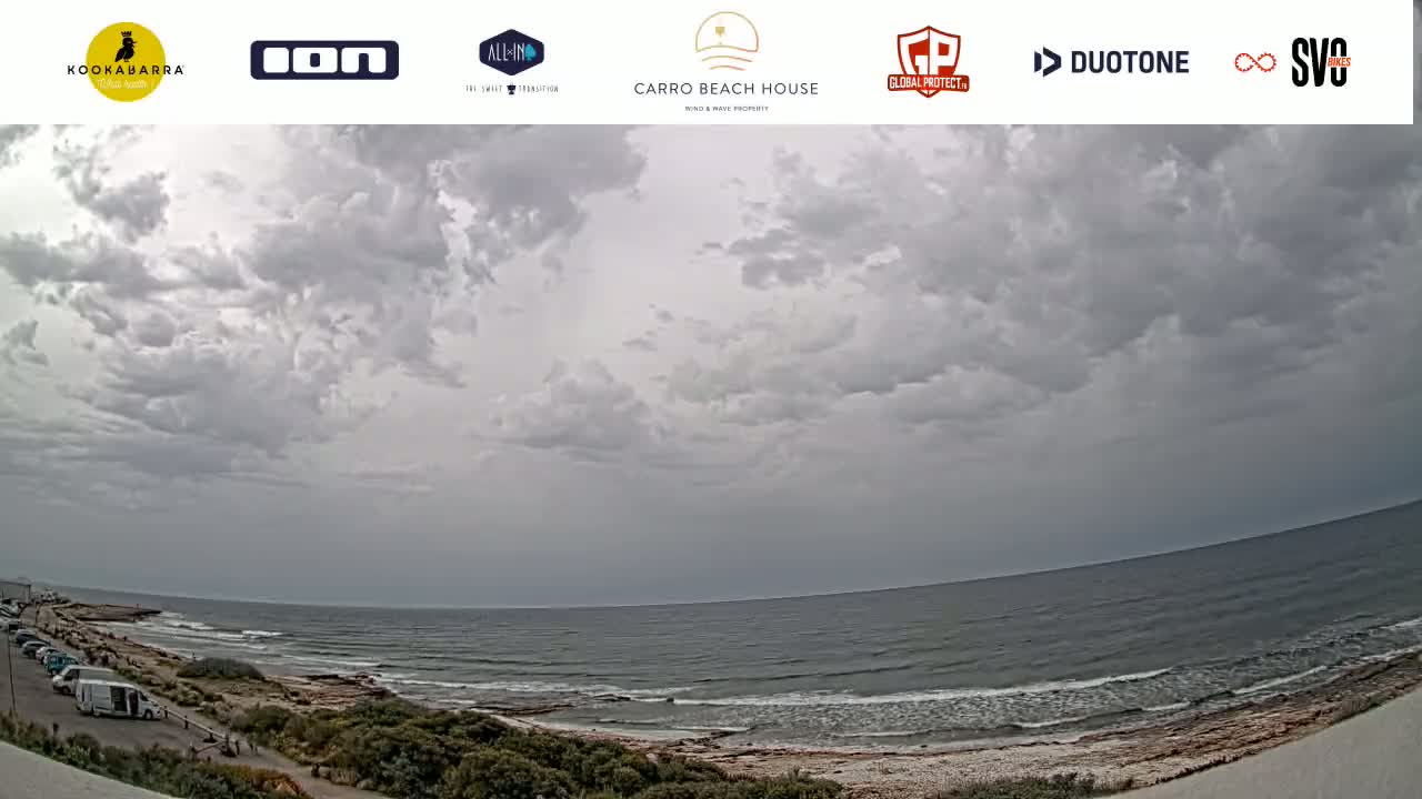 A mostly cloudy sky hangs over a calm ocean, with a rocky shoreline and a parking lot visible in the foreground.