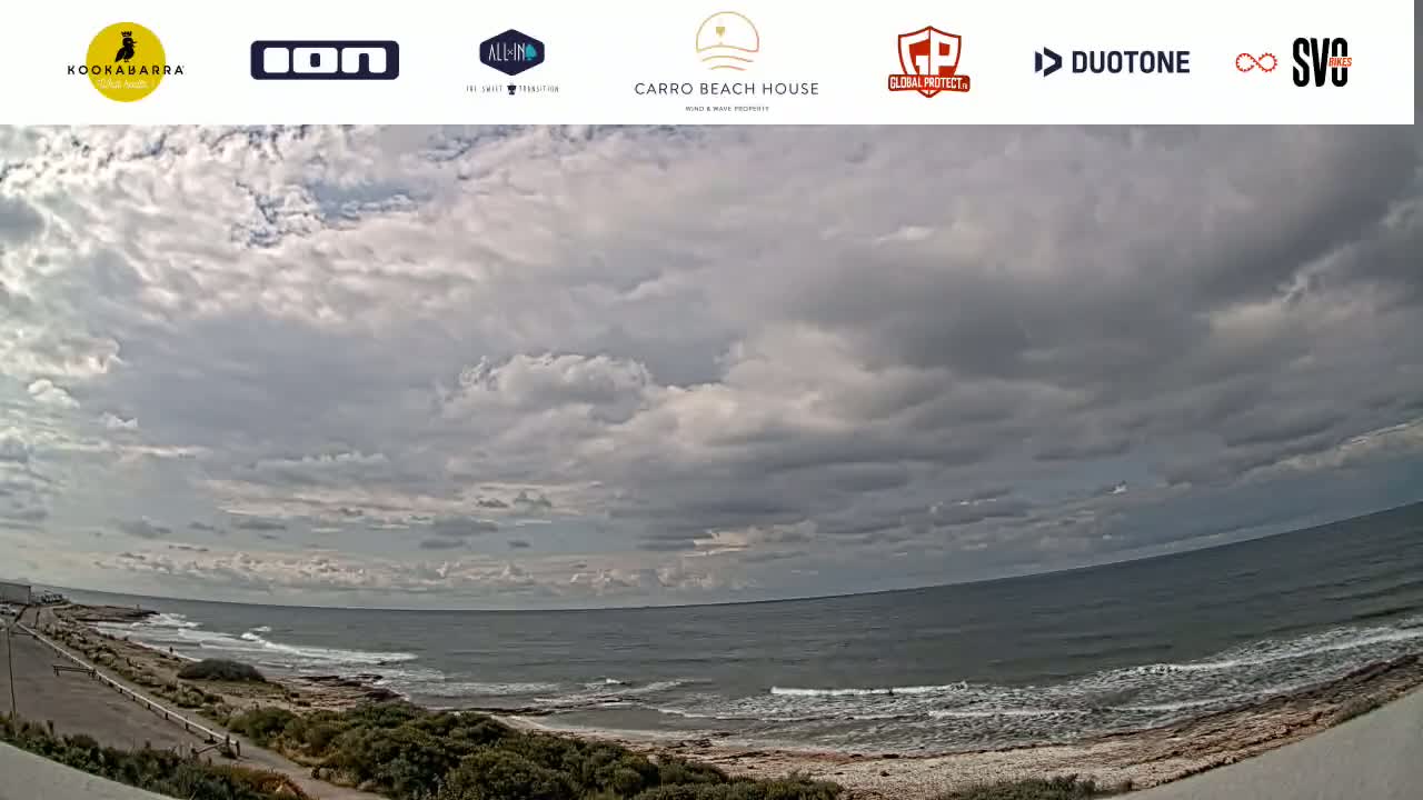A mostly cloudy sky overlooks a rocky coastline with a calm ocean, small waves lapping the shore.