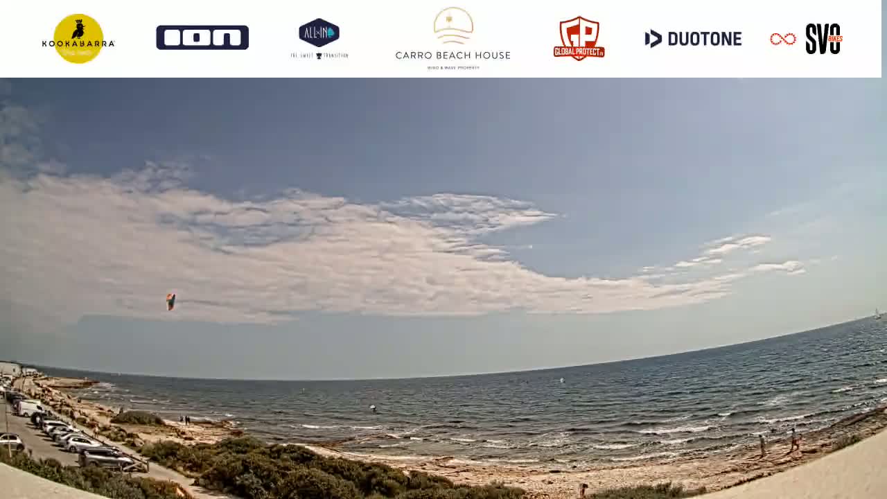 A kite surfer is visible in the distance over a calm sea under a partly cloudy sky.