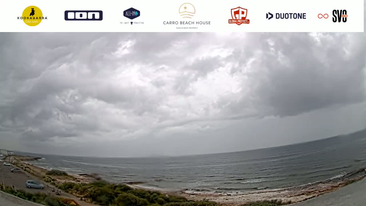 A wide shot shows a cloudy, overcast day at a beach with a calm ocean and a few cars parked along the shore.