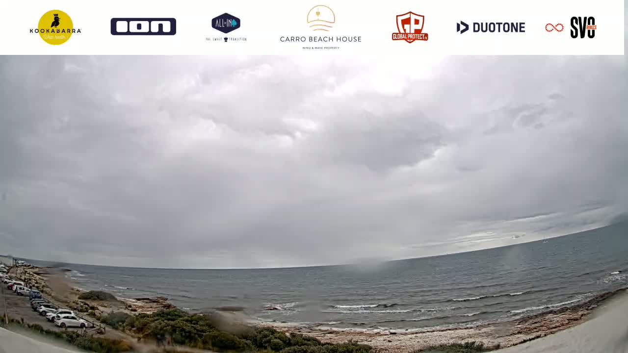 A gray, overcast sky hangs over a calm ocean meeting a sandy shore with cars parked along the roadside and sparse vegetation.