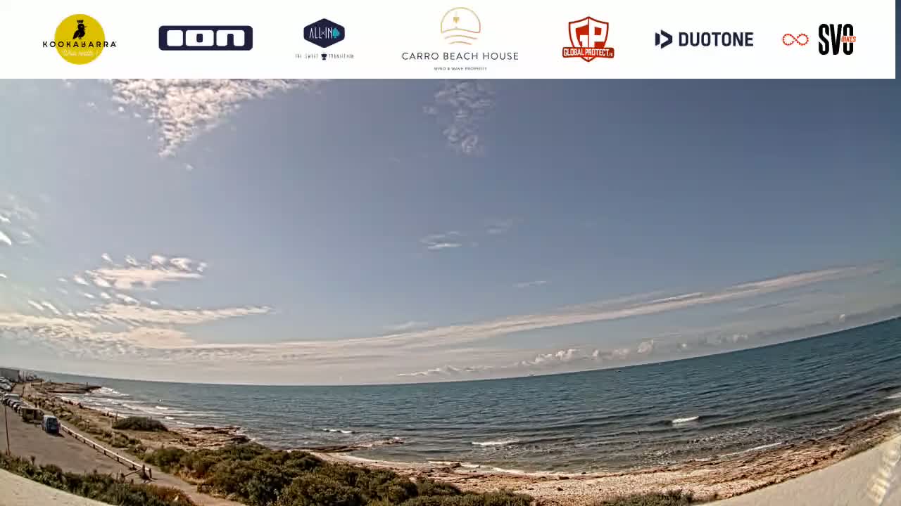 A mostly sunny day reveals a wide, panoramic view of a calm ocean meeting a sandy shore lined with sparse vegetation.
