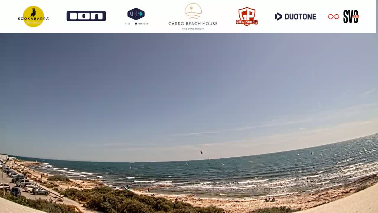A wide, sunny shot shows a sandy beach with a few people and parked cars, next to a calm, blue ocean with windsurfers and kitesurfers.