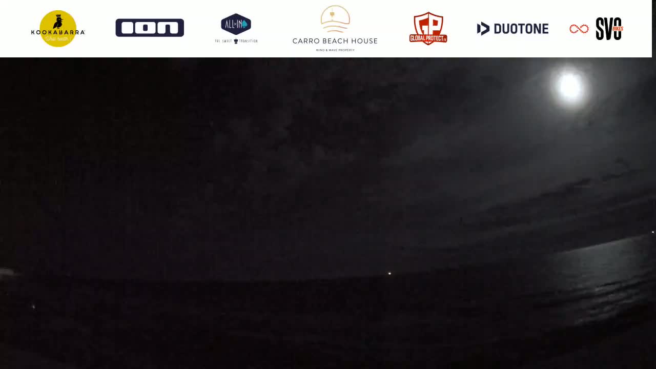 A bright moon illuminates a dark, nighttime beach and ocean under a cloudy sky.