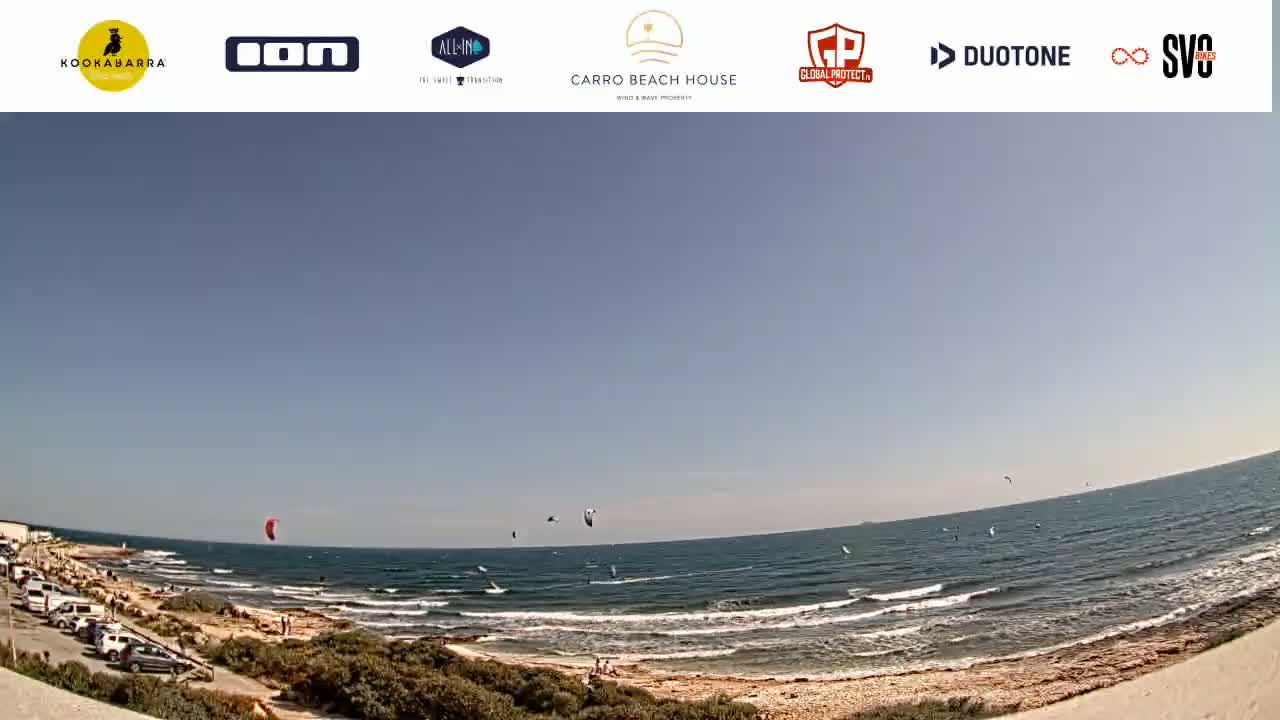 A wide shot of a beach with several kitesurfers in the ocean under a clear, sunny sky.