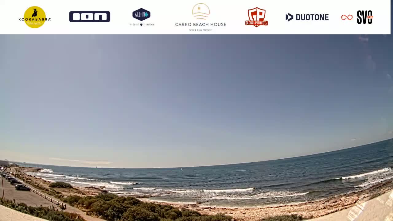 A mostly sunny day at a beach with calm waves lapping a sandy shore, and vegetation bordering the parking lot.