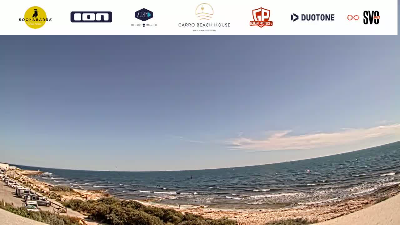 A mostly sunny day at a beach with calm waves, a sandy shoreline, and a parking lot with several cars.