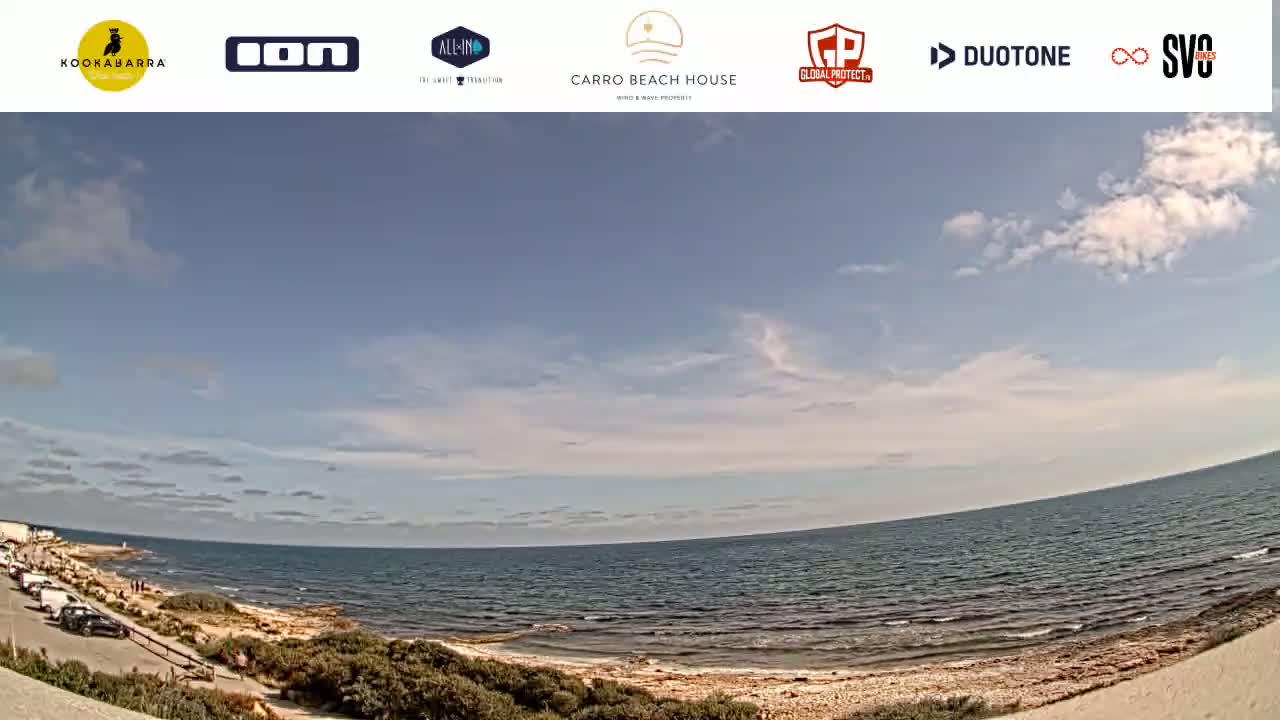 A mostly sunny day at a beach with a calm sea and a few people near the shore.