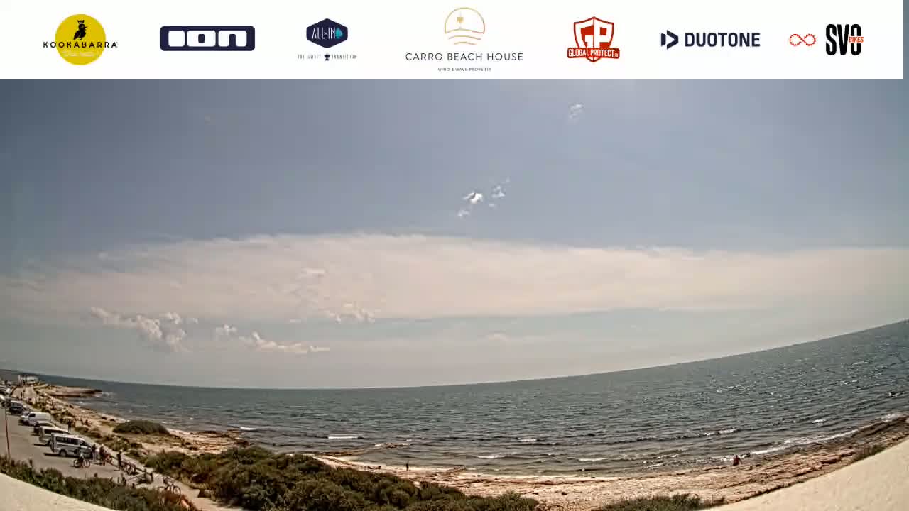 A partly cloudy day shows a wide view of a calm ocean meeting a sandy shoreline with sparse vegetation and a few people visible.