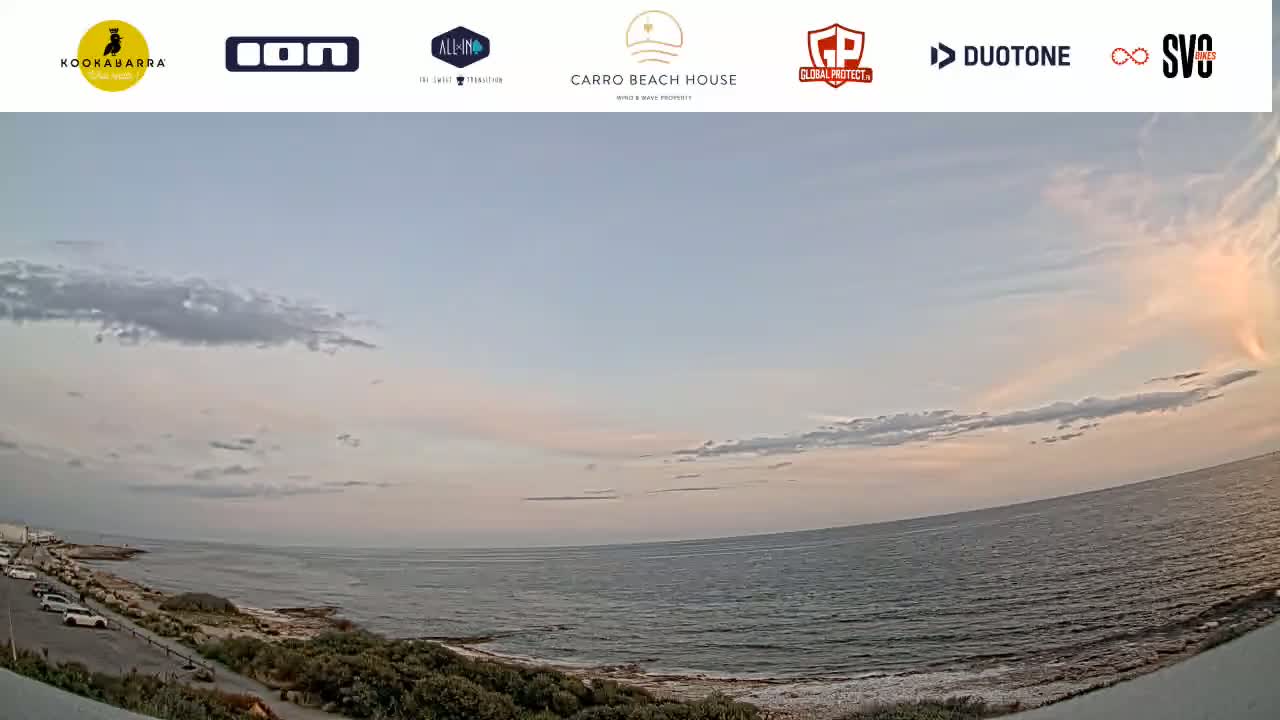 A wide, panoramic view shows a calm ocean under a partly cloudy, pale sunset sky, with a coastal road and vegetation visible in the foreground.