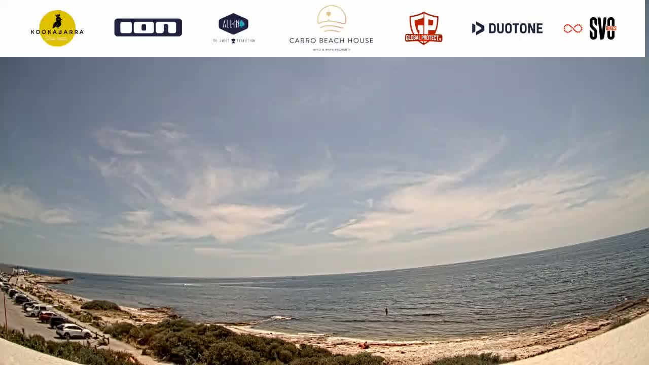 A partly cloudy day at a beach with a rocky shoreline, a calm ocean, and a parking lot with cars visible.