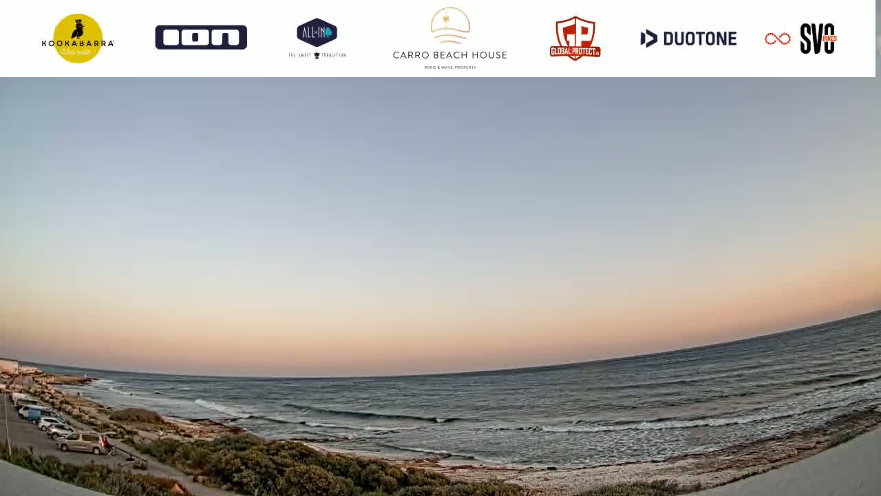 A wide, panoramic view shows a calm ocean meeting a sandy shore at sunset, with several cars parked near the water's edge.