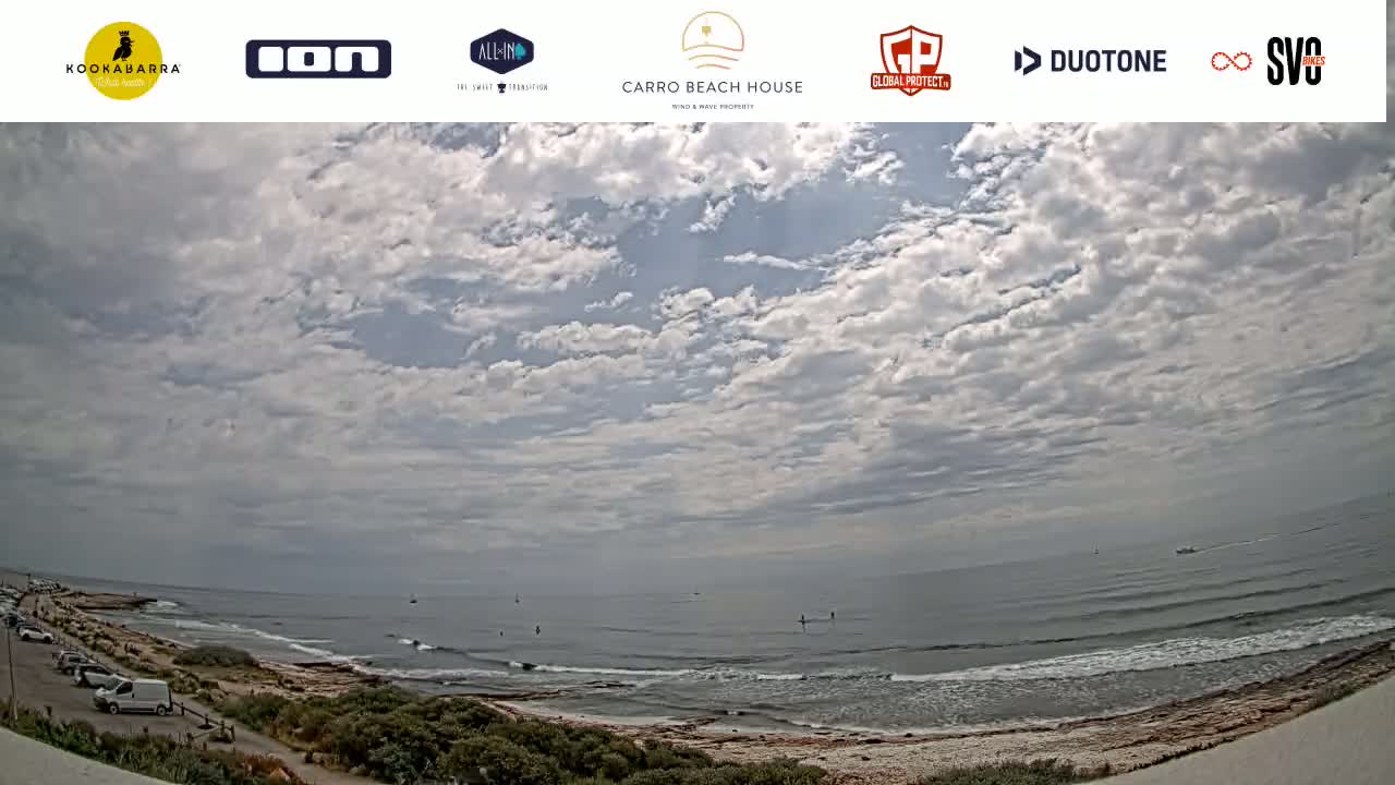 A mostly cloudy sky overlooks a calm ocean with a sandy shoreline and a few cars parked near the water.