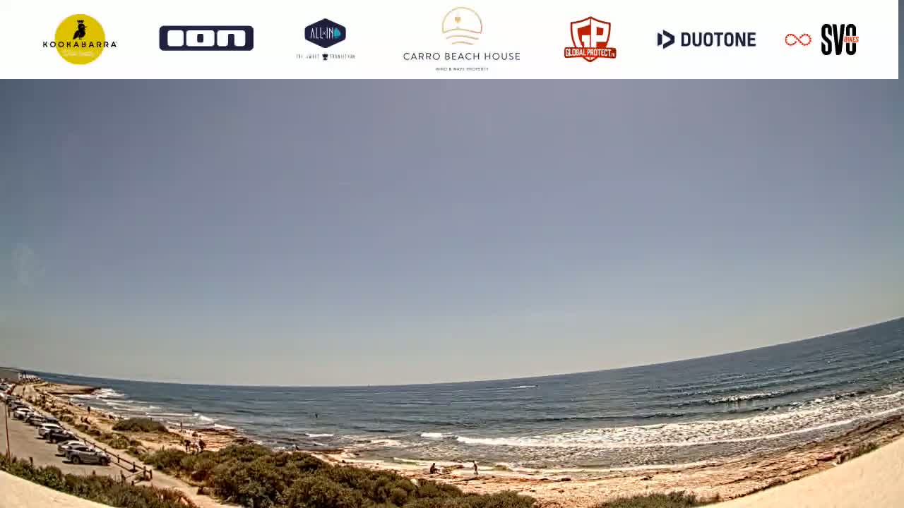 A sunny day at a beach shows a calm ocean with small waves lapping a sandy shore, where a few people are present and cars are parked nearby.