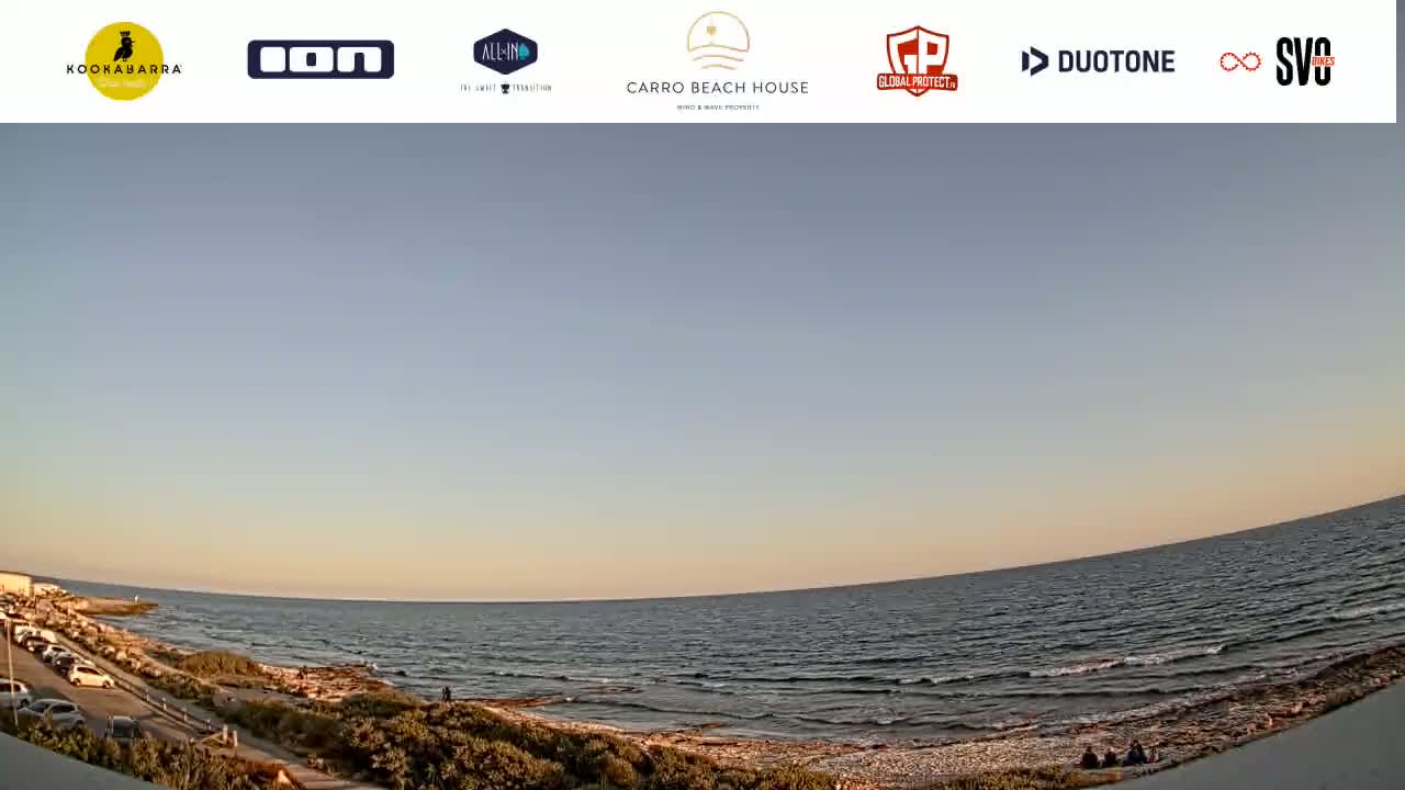 A wide, panoramic view of a calm ocean under a clear, pale-blue sky at sunset, showing a sandy beach with a few people near the water's edge and a parking lot with cars in the distance.