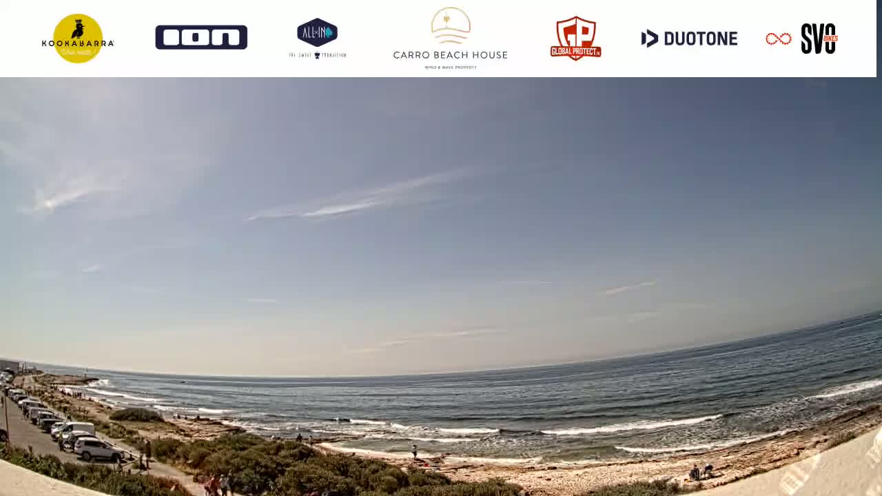 A mostly sunny day at a beach with a sandy shore, calm ocean waves, and a parking lot with several cars visible near the coastline.