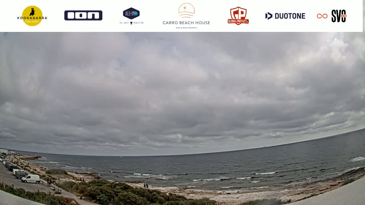 A wide shot of a rocky beach and ocean under a cloudy sky shows a few people on the beach and cars in a parking lot.