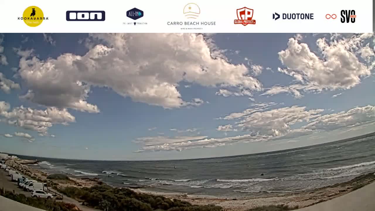 A partly cloudy day shows a wide expanse of ocean with small waves lapping a sandy shore, and cars parked along a road next to sparse vegetation.