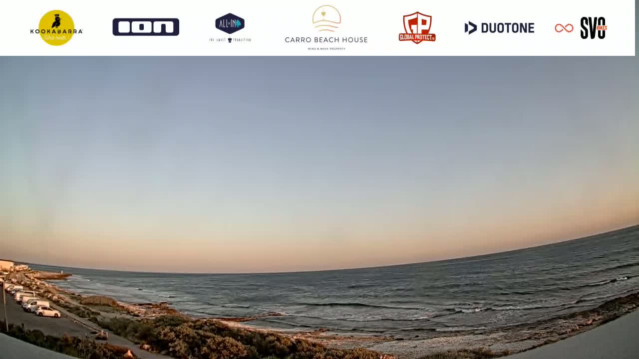 A calm ocean with small waves meets a sandy shore under a pale sunset sky.