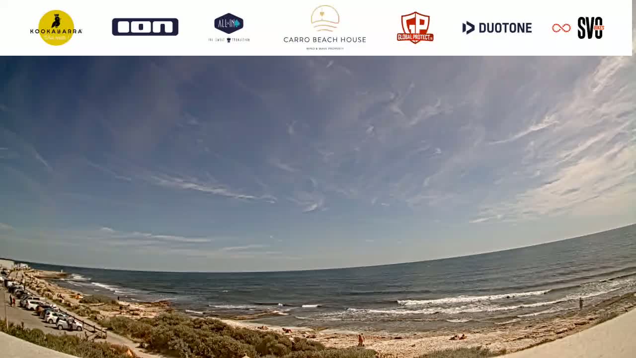 A panoramic view of a beach with a calm ocean under a mostly clear sky with scattered cirrus clouds.