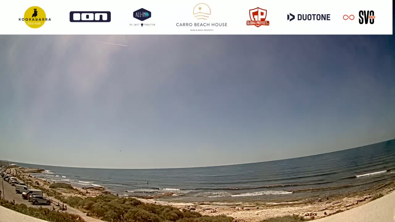 A mostly sunny day at a beach with calm waves, a sandy shore, and a parking lot with several cars.