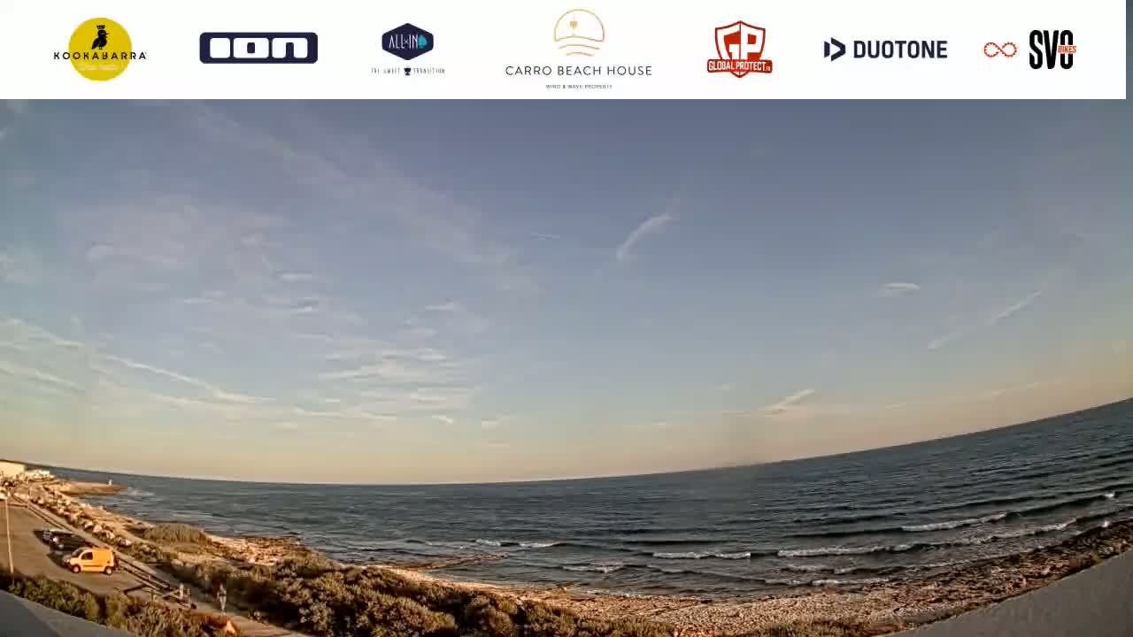 A mostly sunny day overlooks a calm ocean with small waves lapping a rocky shore, and a few people are visible along a road next to the water.