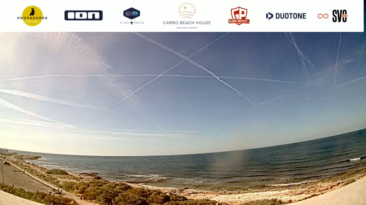 A wide, panoramic view shows a calm ocean under a mostly clear sky with several contrails crossing.