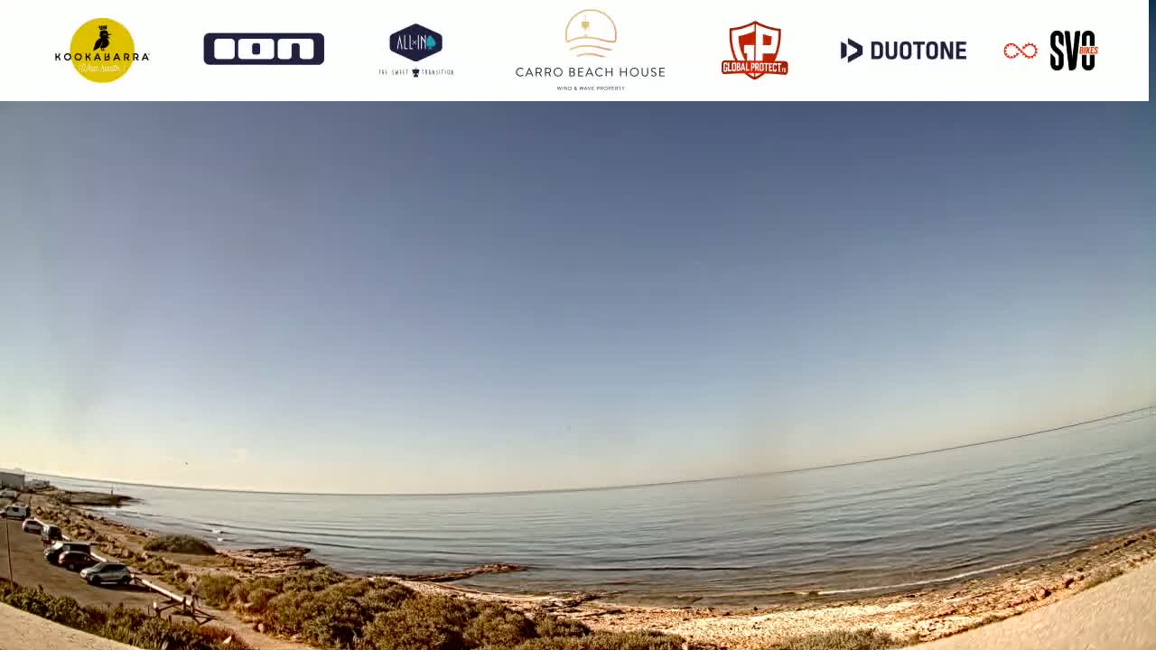 A calm, clear day unfolds at a beach with a rocky shoreline and a few cars parked nearby.