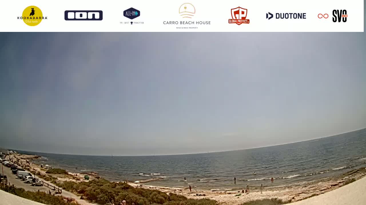 A wide shot of a beach on a sunny day shows a calm ocean, a sandy shore with a few people, and a parking area with cars.