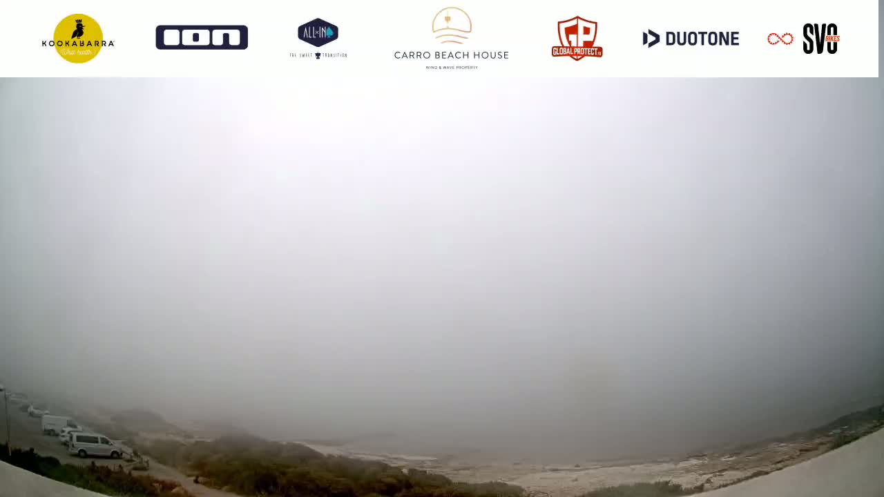 A foggy coastal scene shows a parking lot with a few vehicles near low vegetation, overlooking a barely visible beach in the distance.
