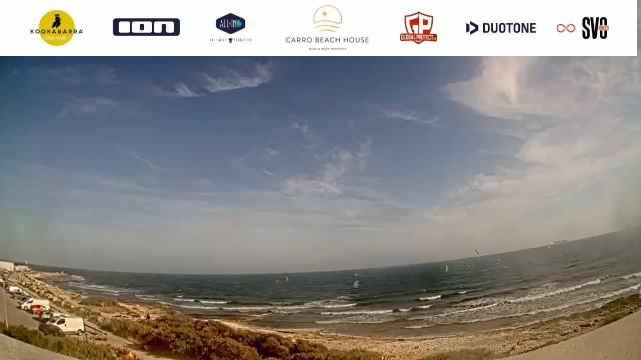 A mostly sunny day with some scattered clouds overlooks a beach with a calm sea and several windsurfers visible in the distance.