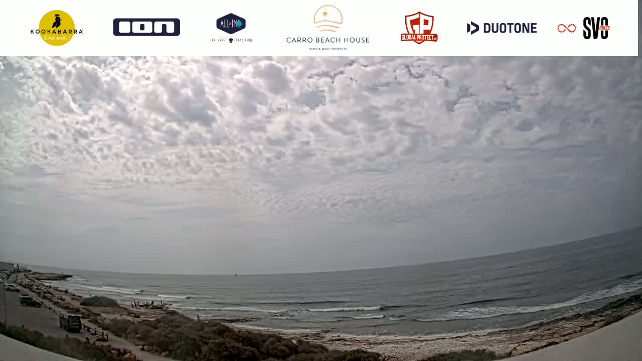 A wide shot shows a partly cloudy sky over a calm ocean, with a beach and a road visible in the foreground.