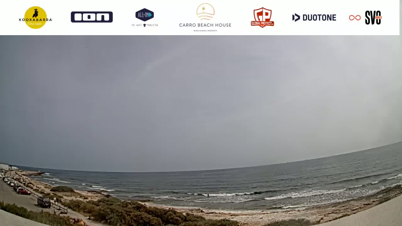 A wide, low-angle shot shows a mostly cloudy day at a beach with small waves lapping the shore, a few vehicles parked in a lot, and sparse vegetation along the sand.