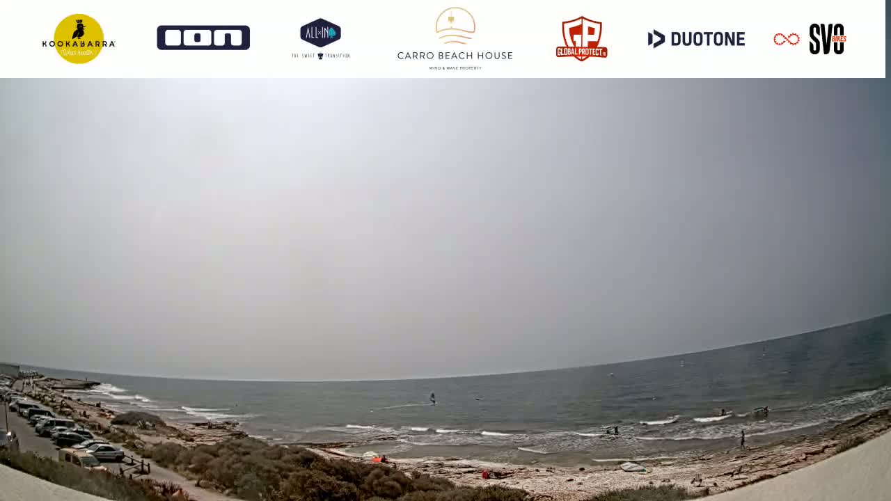 A hazy, sunny day at a beach shows a calm sea with a few people and windsurfers, along with cars parked in a lot overlooking the water.