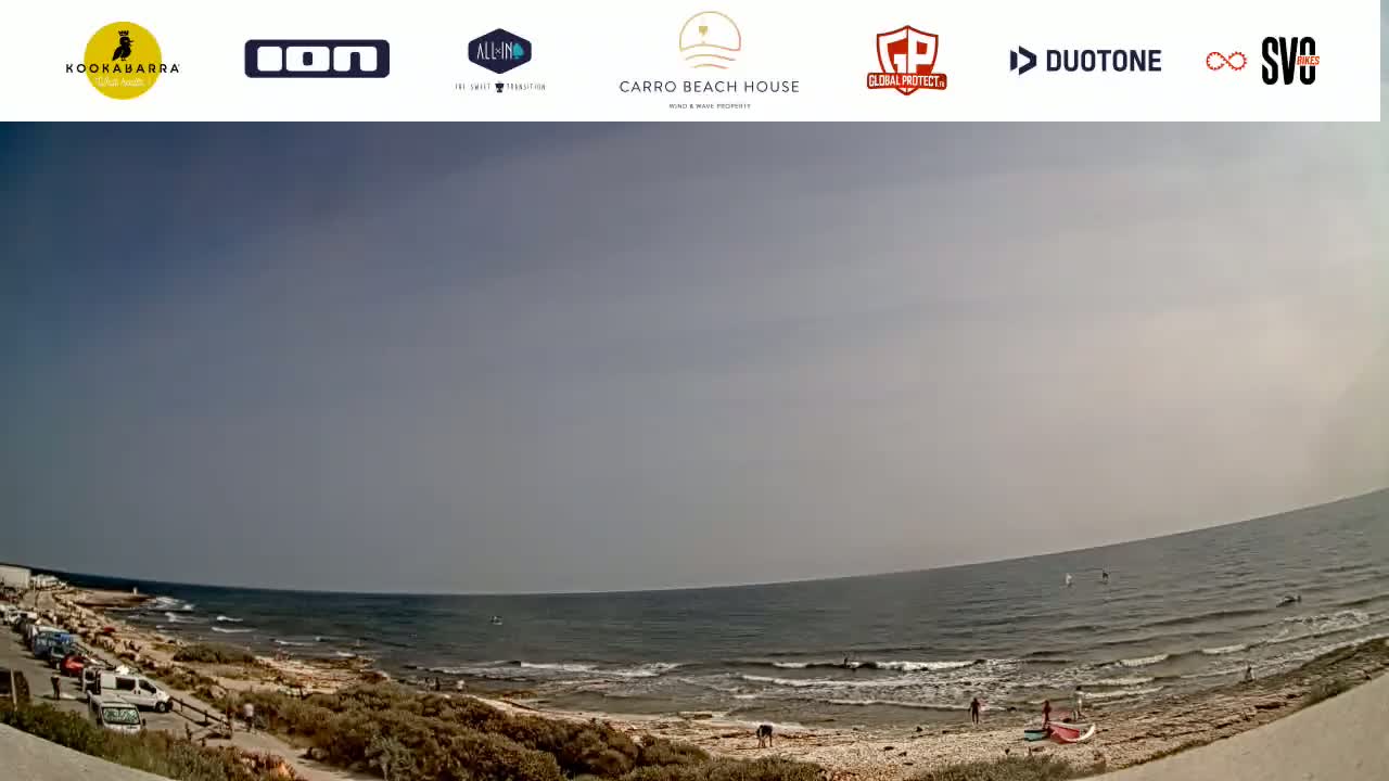 A wide shot shows a mostly calm ocean under a hazy sky, with a sandy beach and some people visible along the shoreline and in the water.