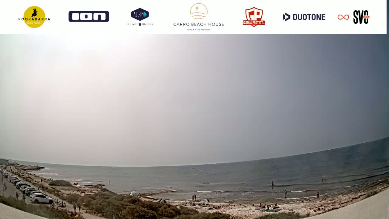 A hazy, sunny day at a beach with calm water, a sandy shore, and a few people scattered along the coast and in the water, with cars parked along a road nearby.