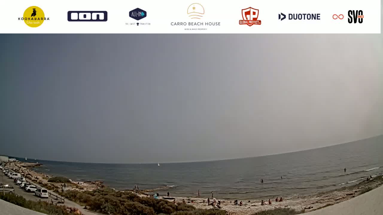 A hazy, sunny day at a beach shows a calm sea with people on the shore and parked cars nearby.