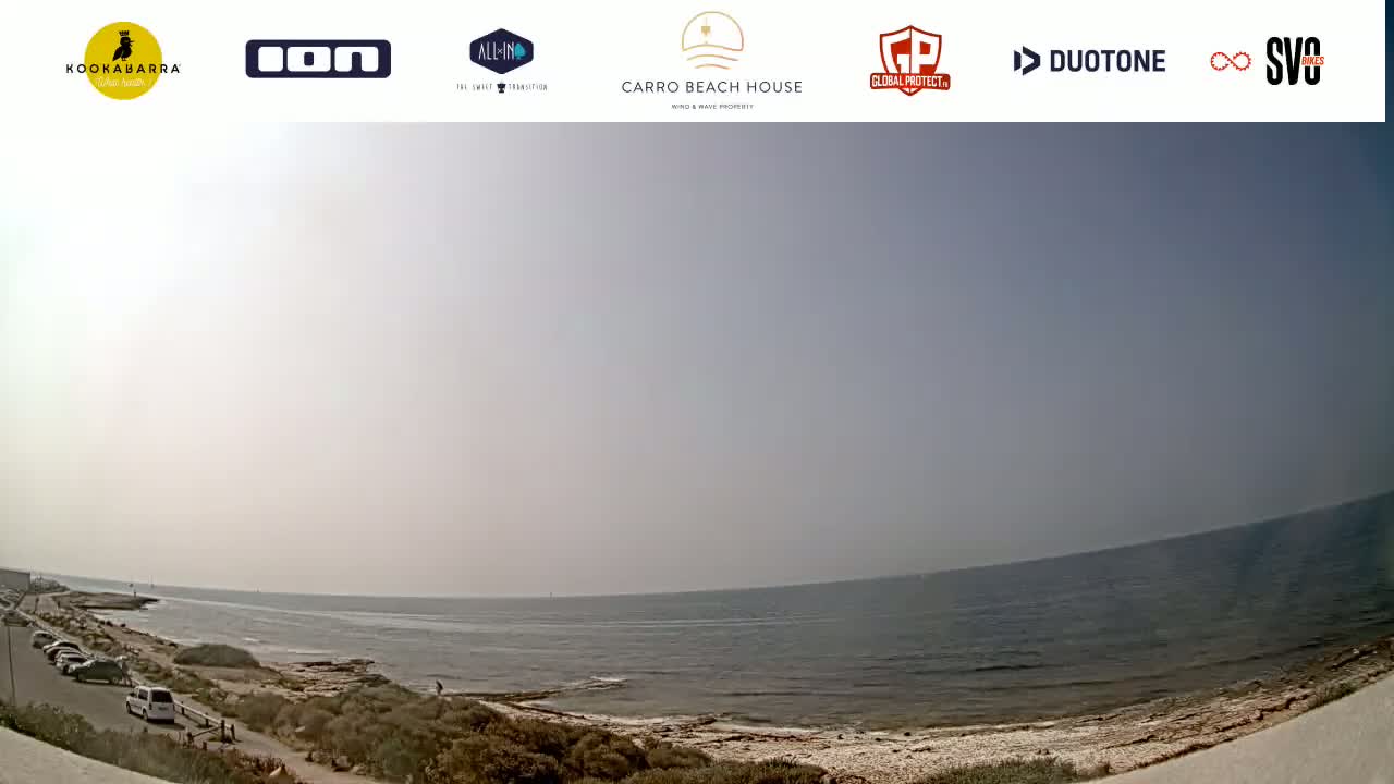 A wide, panoramic view shows a calm ocean under a hazy sky, with a small parking lot and vegetation along the shoreline.