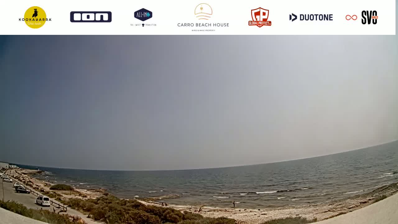A mostly sunny day shows a calm sea lapping a rocky shoreline with sparse vegetation and a few parked cars visible in the distance.