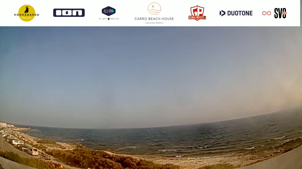 A wide shot shows a calm sea with small waves lapping a sandy shore, under a hazy sky.