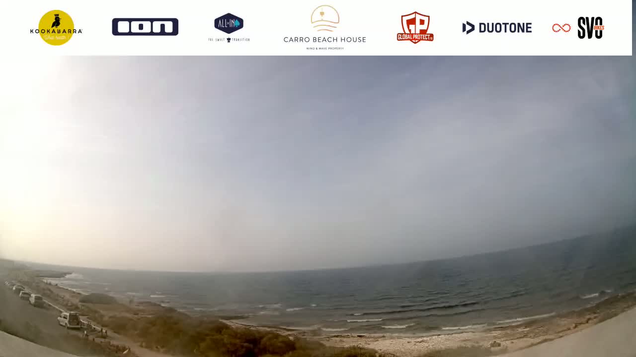 A hazy, overcast day at a beach shows a calm sea with a sandy shore and a few vehicles parked near the water.