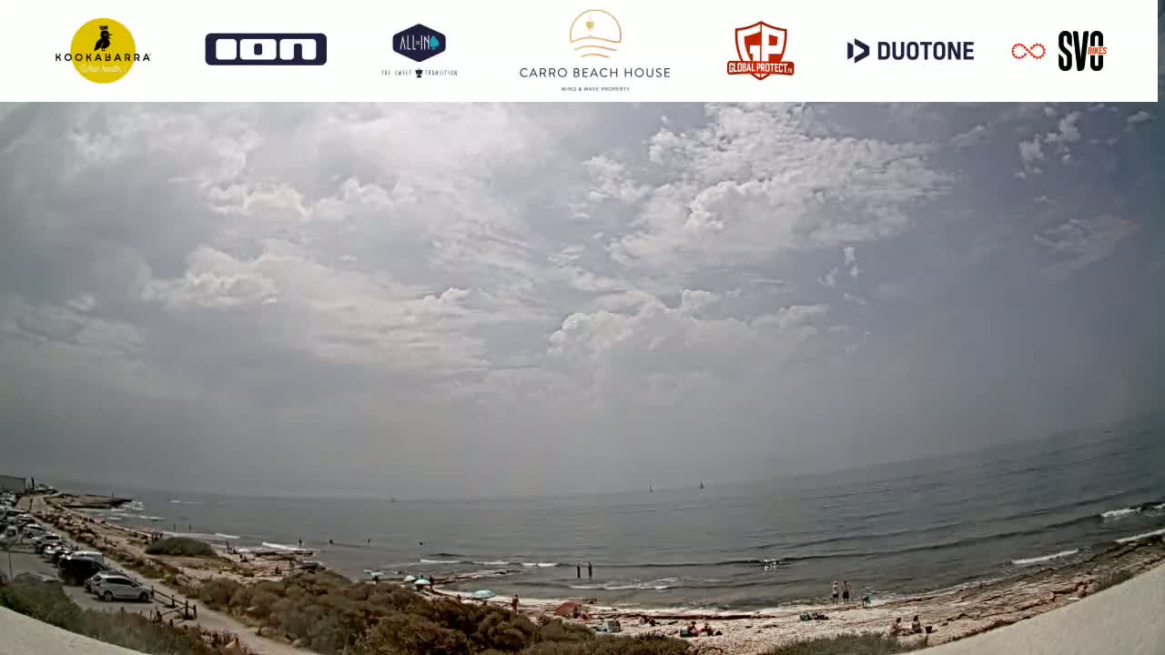 A partly cloudy day at a beach with people swimming and relaxing on the shore and parked cars visible in a parking lot.