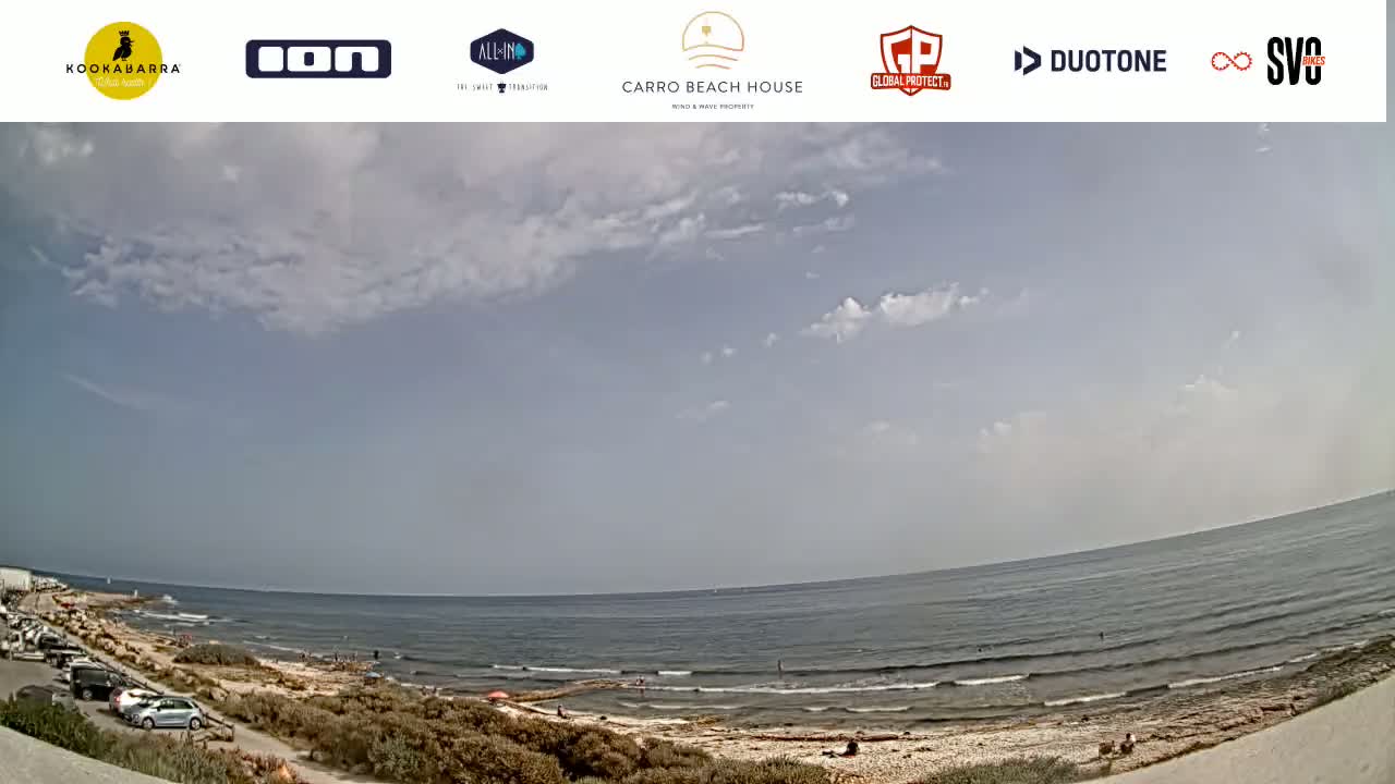 A partly cloudy day at a beach shows a calm ocean with a few people in the water and cars parked near the sandy shore.