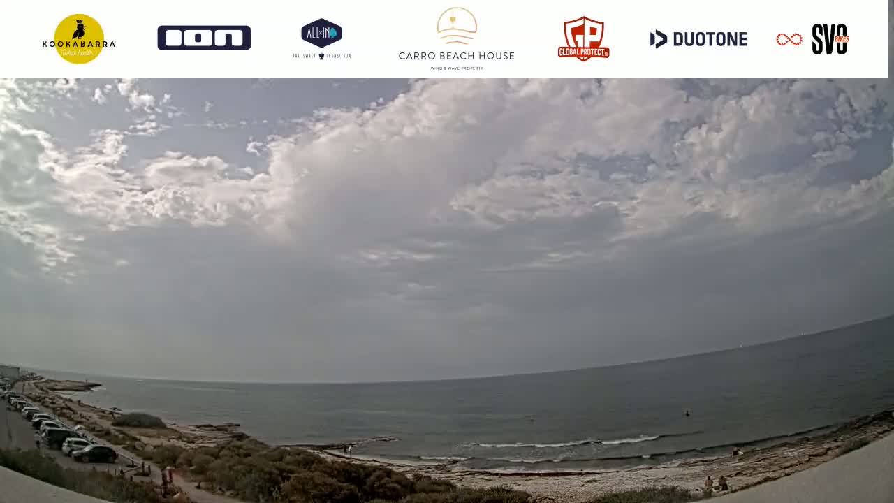 A partly cloudy day overlooks a calm ocean with a rocky shore and a parking lot of cars visible near the water.