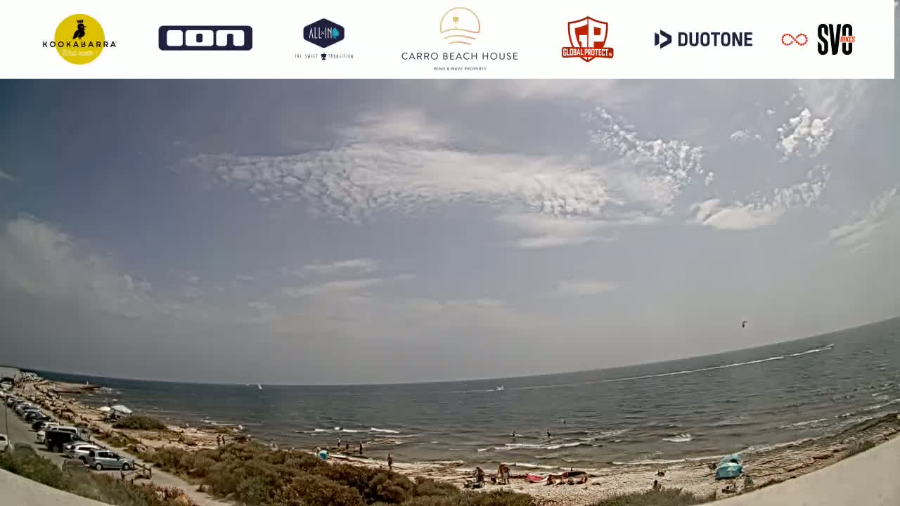 A partly cloudy day at a beach shows a calm sea with a few people on the shore and a kite surfer in the distance.