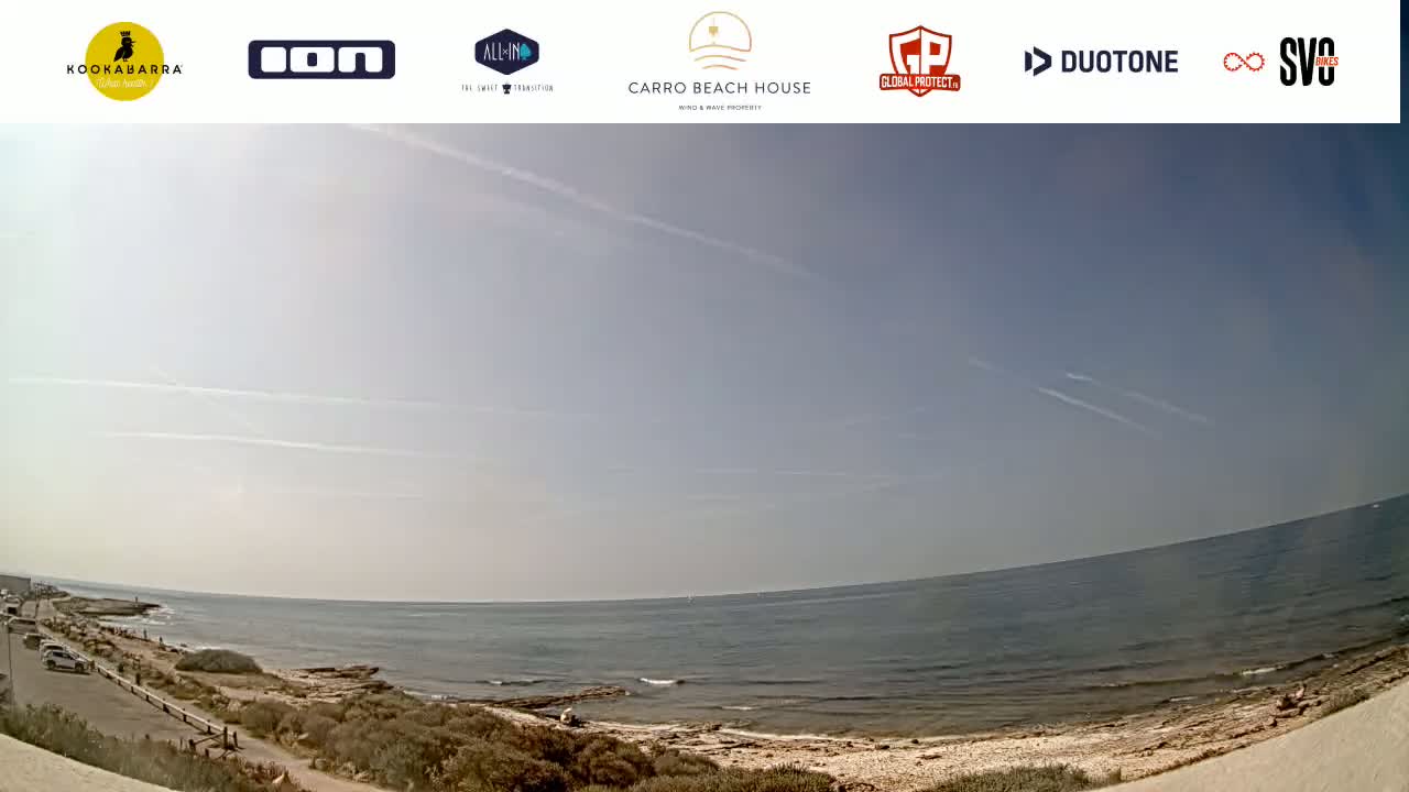 A mostly sunny day shows a wide, sandy beach and calm ocean, with a few people visible in the distance.